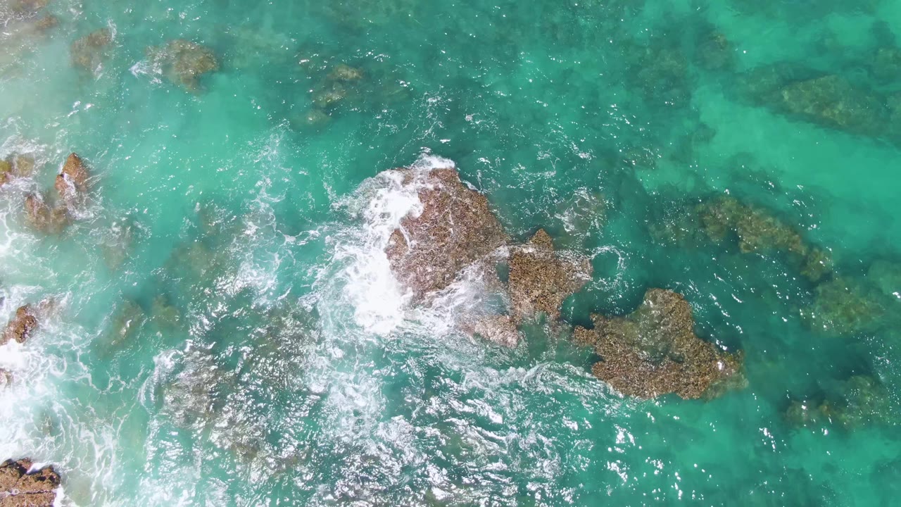 Overhead frame from Buzzards Beach, Baja California Sur, with surf wrapping the central rock cluster and darker submerged seabed visible beneath the clear turquoise water, with no visible people.