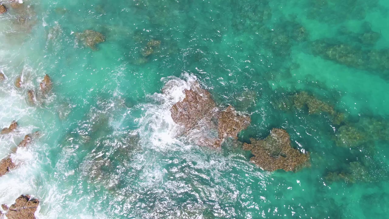 Buzzards Beach, Baja California Sur appears from above with a large foamy break at left, a central exposed rock, and submerged rock patches under turquoise water, with no visible people.