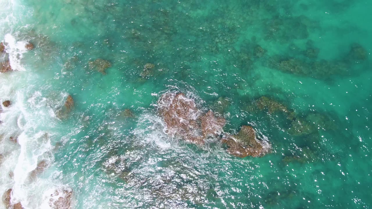 Top-down view of turquoise water at Buzzards Beach, Baja California Sur, with white surf pushing in from the left, brown rocks near center, and no visible people.