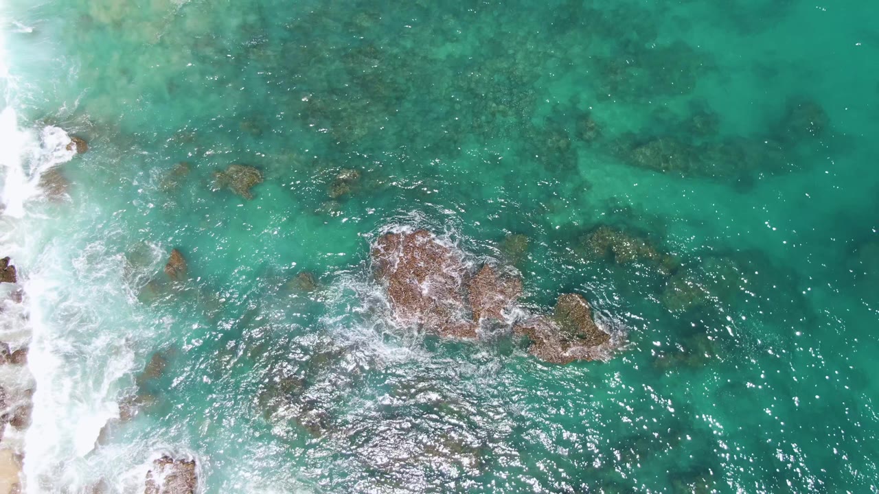 Aerial frame over Buzzards Beach shows white foam curling around the central rock cluster, darker submerged patches above it, and no visible people.