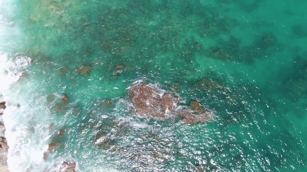 Top-down drone view at Buzzards Beach captures surf entering from the left edge, a line of brown rocks across clear green-blue water, and no visible people.