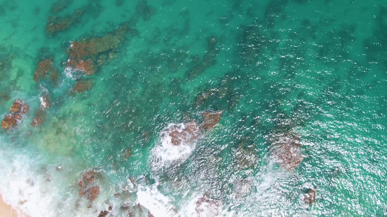 At Buzzards Beach in Baja California Sur, clear blue-green water surrounds rust-colored rock clusters while white foam spreads along the lower edge, with no visible people.