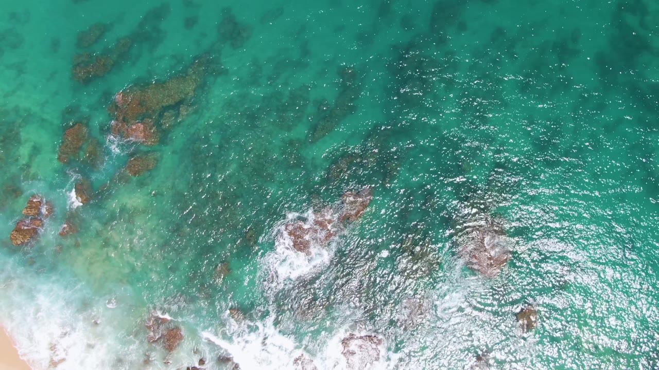 At Buzzards Beach in Baja California Sur, scattered rocks sit beneath and above turquoise water as bright foam curls around the lower shoreline, with no visible people.