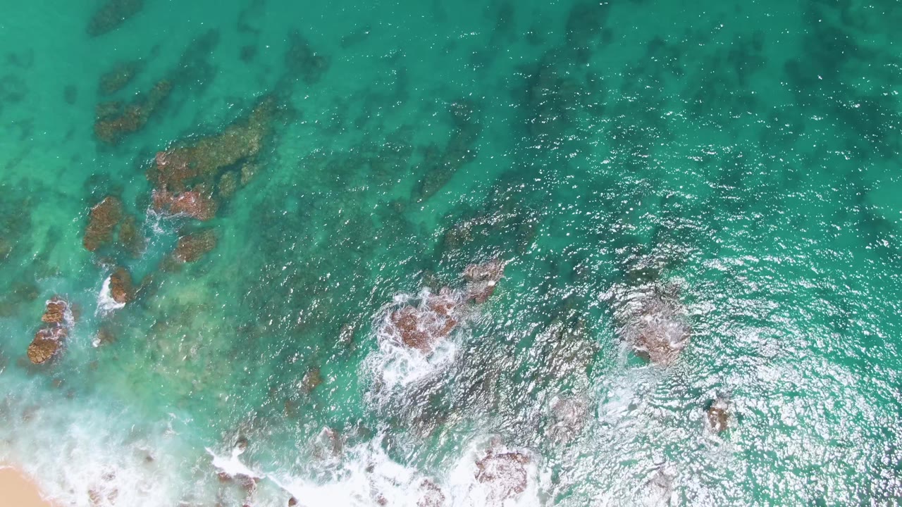 At Buzzards Beach in Baja California Sur, shallow green-blue water reveals dark underwater rocks while a band of white foam crosses the bottom of the frame, with no visible people.