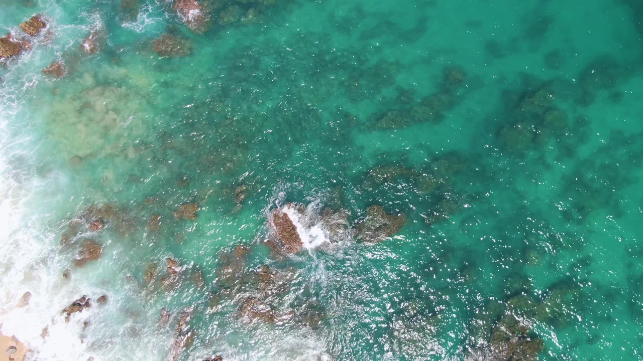 Aerial frame at Buzzards Beach showing pale shallow water on the left, scattered brown rocks under turquoise sea, and foam breaking around a central rock with no visible people.