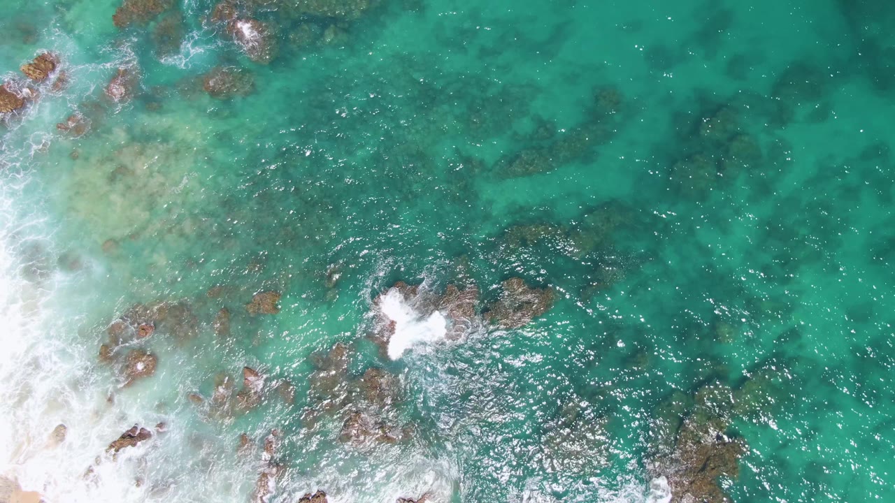 Top-down Buzzards Beach view with deeper turquoise water to the right, rocky patches below the surface, and a compact white foam break around the central outcrop, with no visible people.
