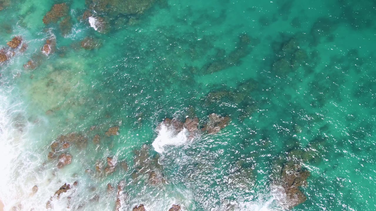 Overhead frame from Buzzards Beach with foamy surf advancing from the left, tan shallow water near the edge, and darker submerged rocks across the turquoise ocean, with no visible people.