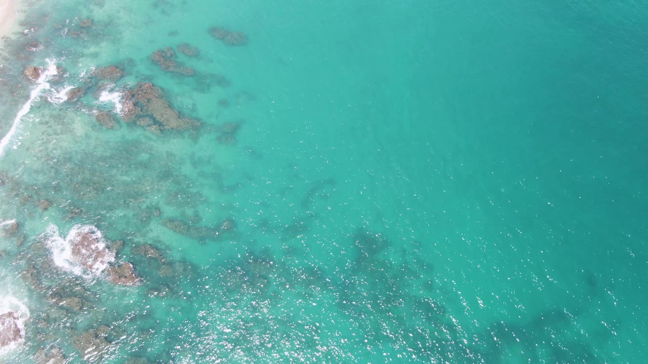 Top-down aerial frame over Buzzards Beach, Mexico, showing turquoise water, submerged rocks, white foam near the left shoreline, and no visible people.