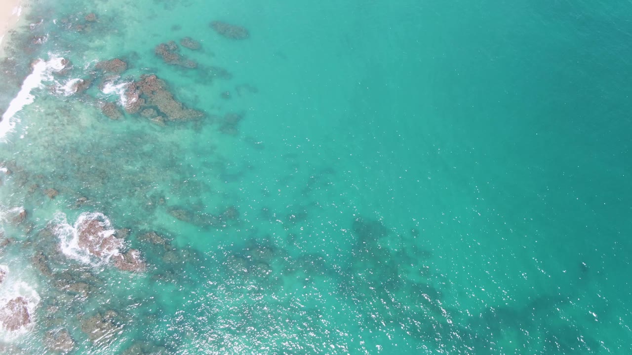 Aerial view at Buzzards Beach, Mexico, with reef rocks clustered left of center, white wave foam curling around them, open turquoise water to the right, and no visible people.