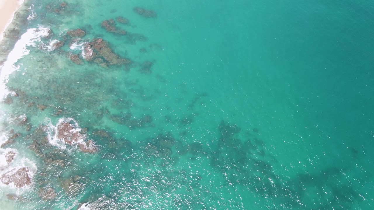 Overhead frame from Buzzards Beach, Mexico, showing the sandy shore along the left edge, scattered rocky shallows, white surf bursts, deeper teal water to the right, and no visible people.
