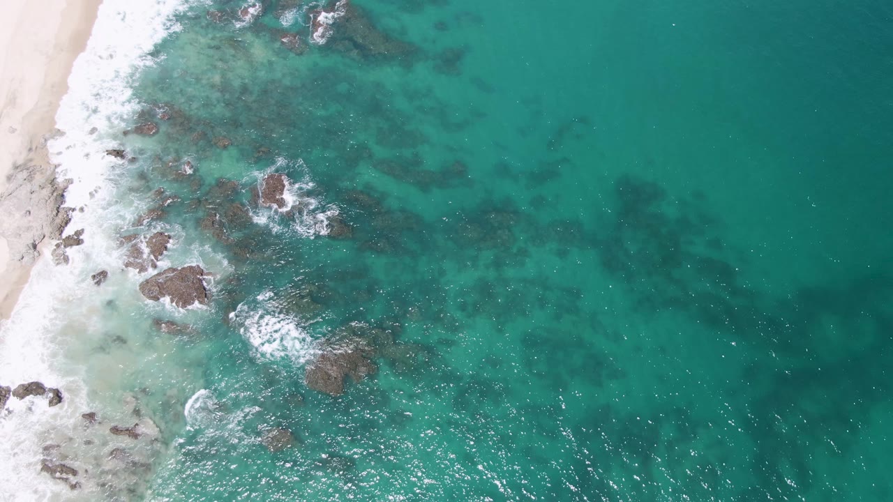 Overhead turquoise water breaking over rocks beside a sandy beach.