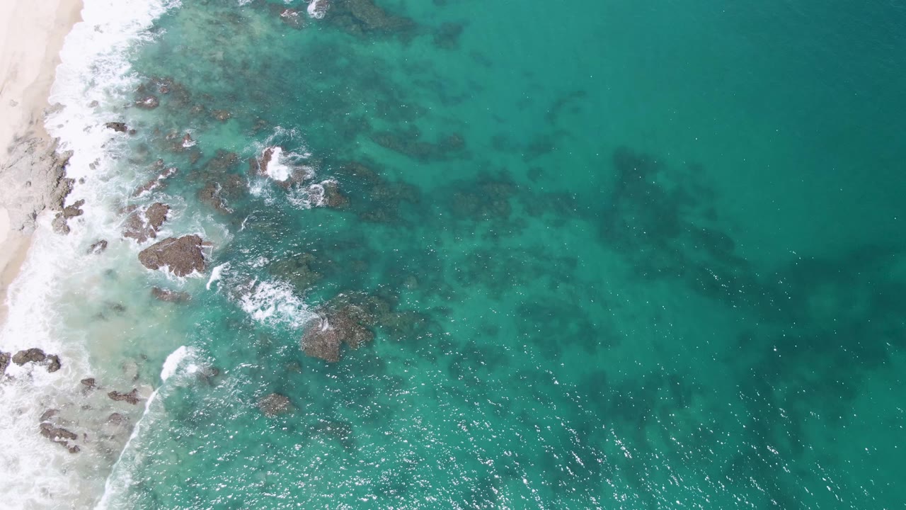 Aerial frame from Buzzards Beach showing foamy wave lines wrapping around exposed and submerged rocks, with pale sand along the left edge and no visible people.