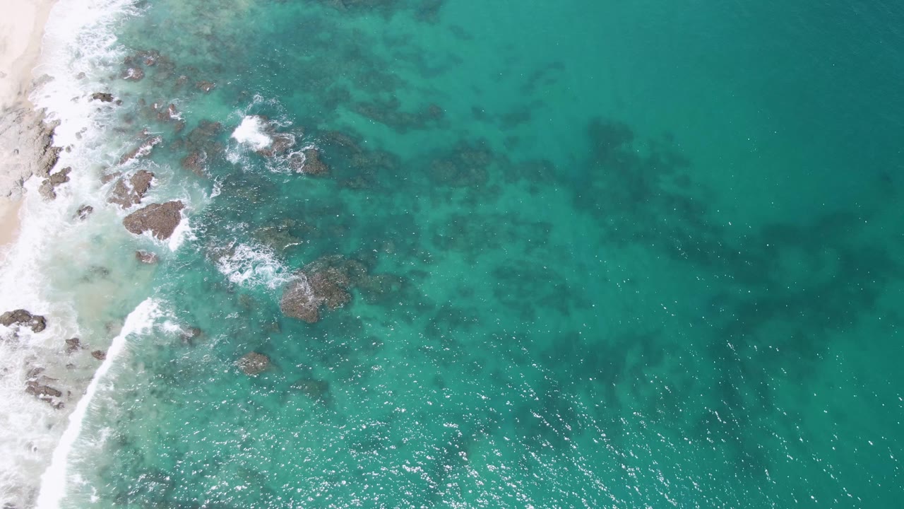 Buzzards Beach appears from above with scattered dark rocks under turquoise water, a narrow sandy edge on the left, bright foam near the rocks, and no visible people.
