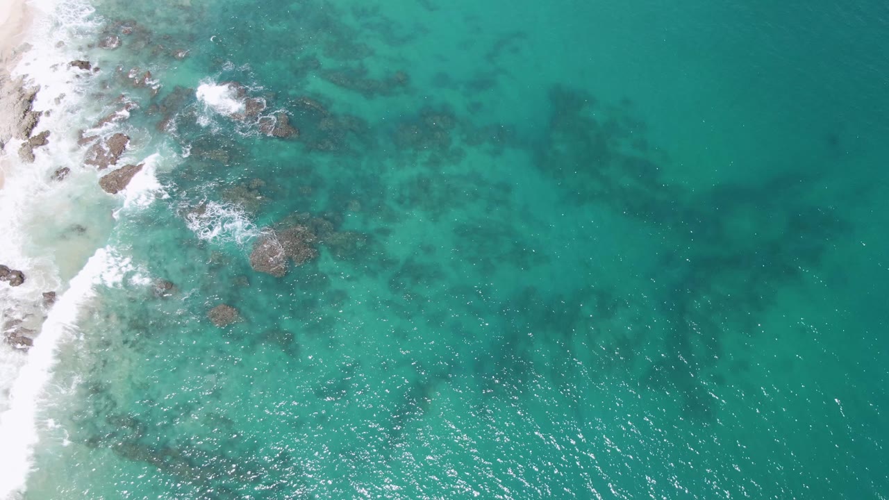 Aerial view at Buzzards Beach with the sand strip along the left border, submerged rocks across the shallows, larger open turquoise water to the right, and no visible people.