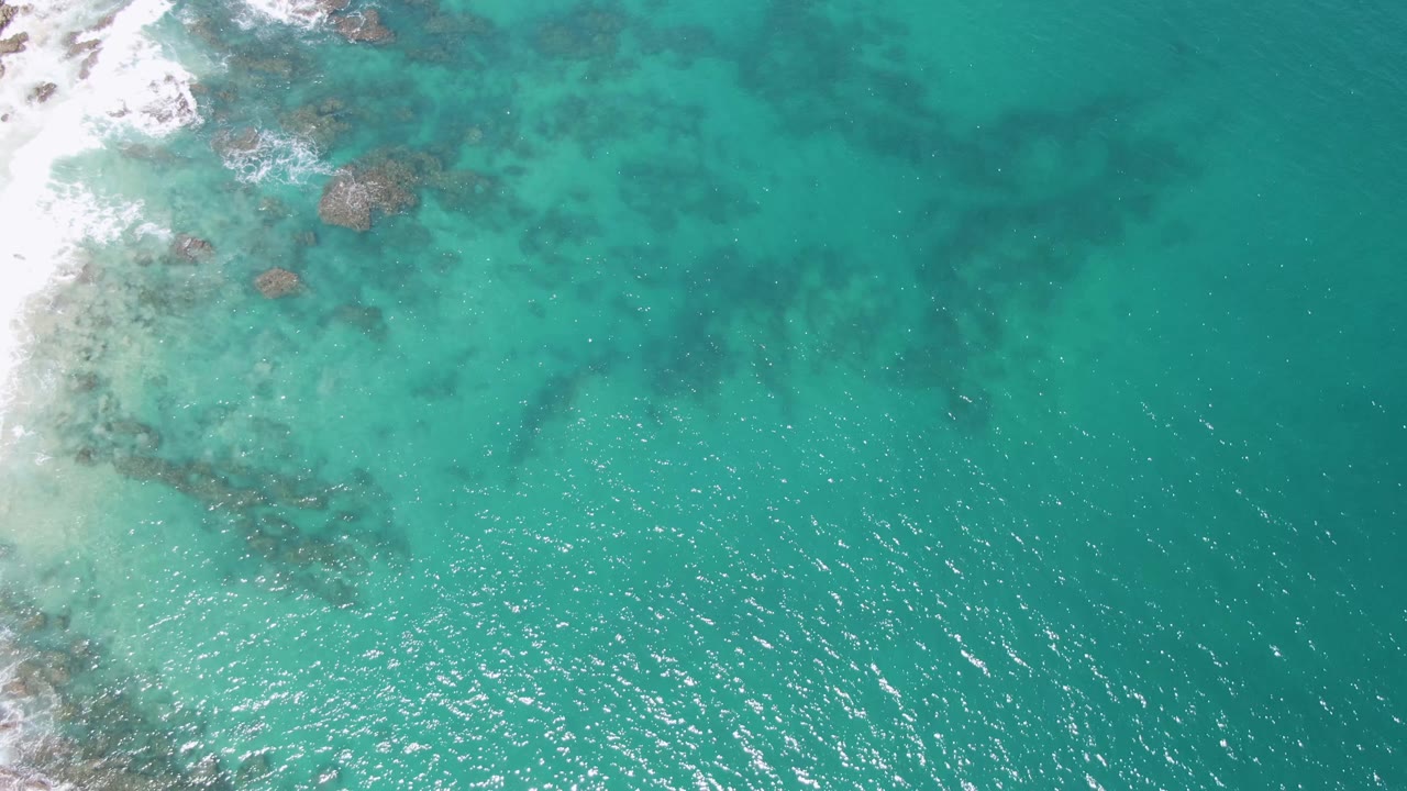 Overhead view of Buzzards Beach turquoise water with dark underwater rocks near the upper left surf line and no visible people.