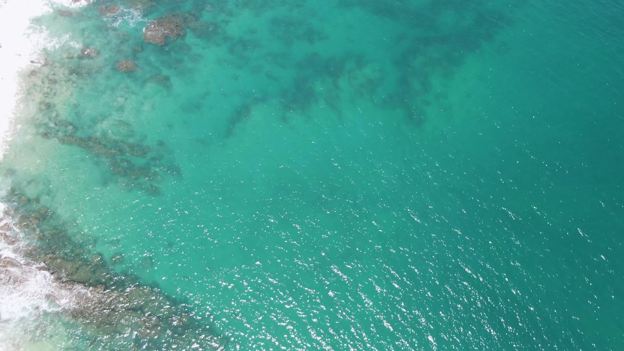 High overhead view of Buzzards Beach with a diagonal rocky surf edge on the left, open turquoise water to the right, and no visible people.