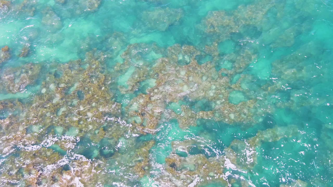 Overhead view at Buzzards Beach, Baja California Sur, showing clear turquoise water over submerged rocks with foam brushing the lower edge and no visible people.
