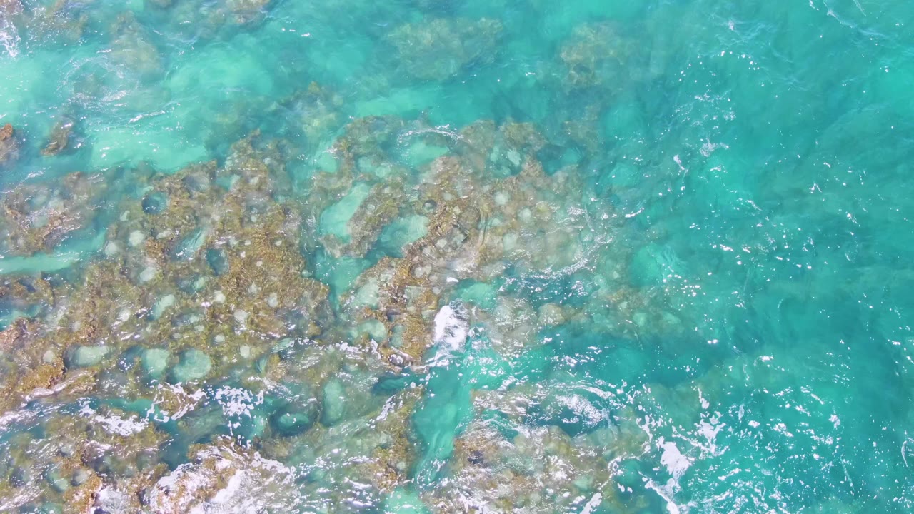 Top-down view at Buzzards Beach with brown and pale submerged rocks under turquoise water, foam gathering near the lower center, and no visible people.