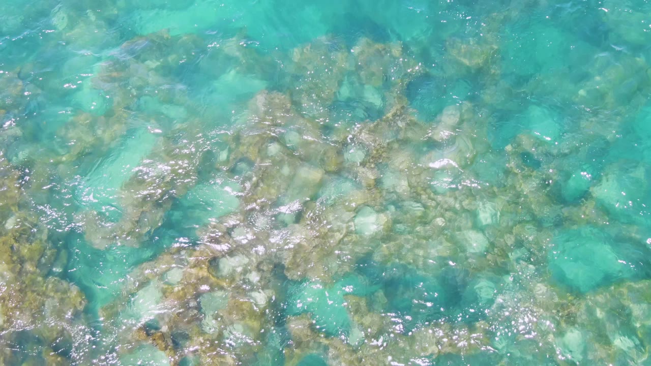 Clear turquoise water ripples over clustered submerged stones at Buzzards Beach, Baja California Sur, with bright surface reflections and no visible people.