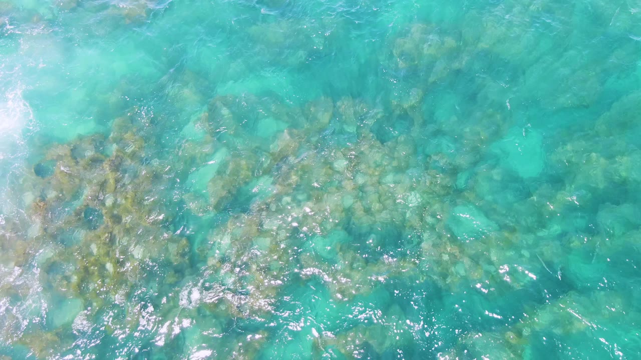 Overhead frame of Buzzards Beach water with tan underwater rock clusters crossing the lower half, turquoise shallows above, bright reflections, and no visible people.