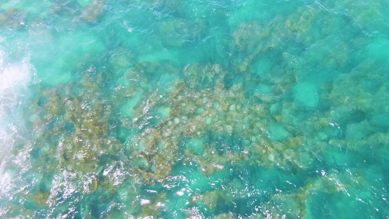 Top-down Buzzards Beach view with the rocky seabed running diagonally through turquoise water, scattered pale stones, moving highlights, and no visible people.