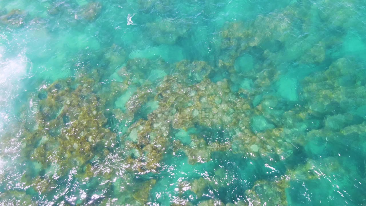 Buzzards Beach shallows seen from above, with a broad rocky patch under clear water, white foam or wave wash entering from the left, and no visible people.