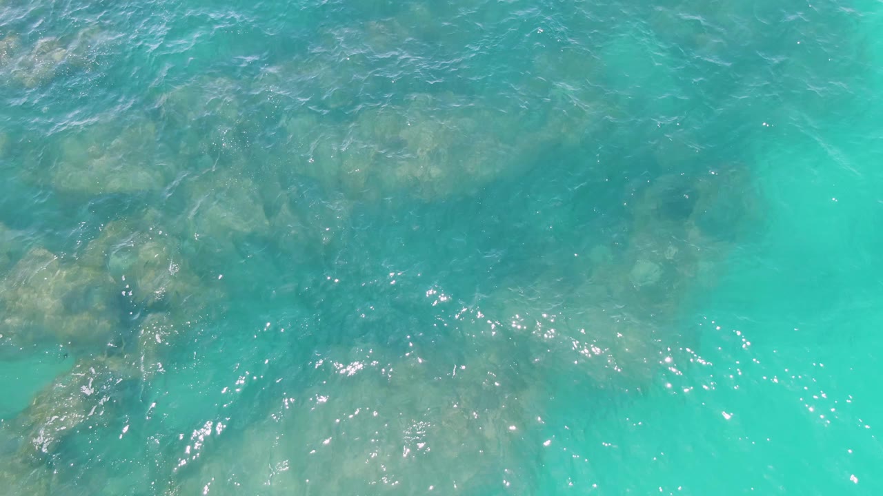 Ripples move across clear turquoise water at Buzzards Beach, with darker submerged rocks clustered toward the left and no visible people.