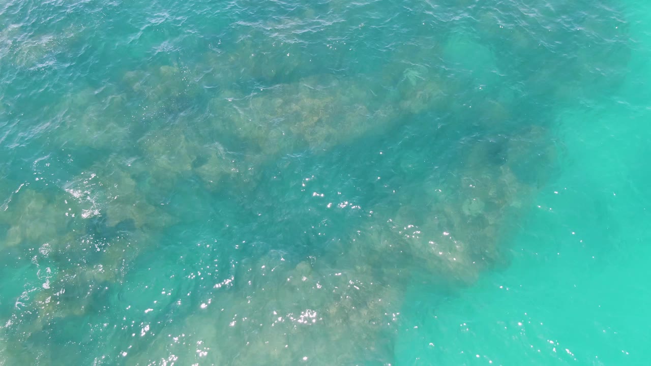 A shallow turquoise ocean view at Buzzards Beach shows underwater rocks across the upper half, scattered sun glints, and no visible people.