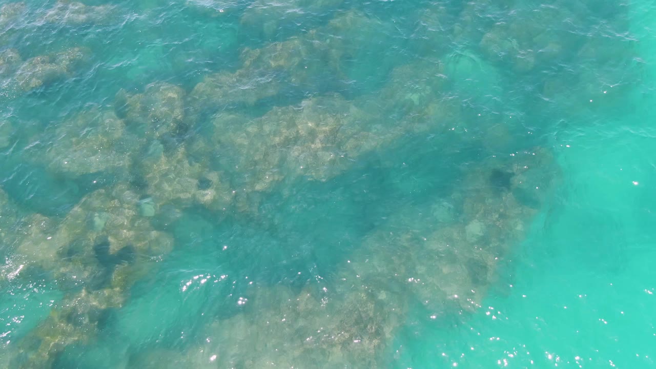 Clear shallow water at Buzzards Beach reveals a broad patch of submerged rocks as ripples shift across the blue-green surface with no visible people.