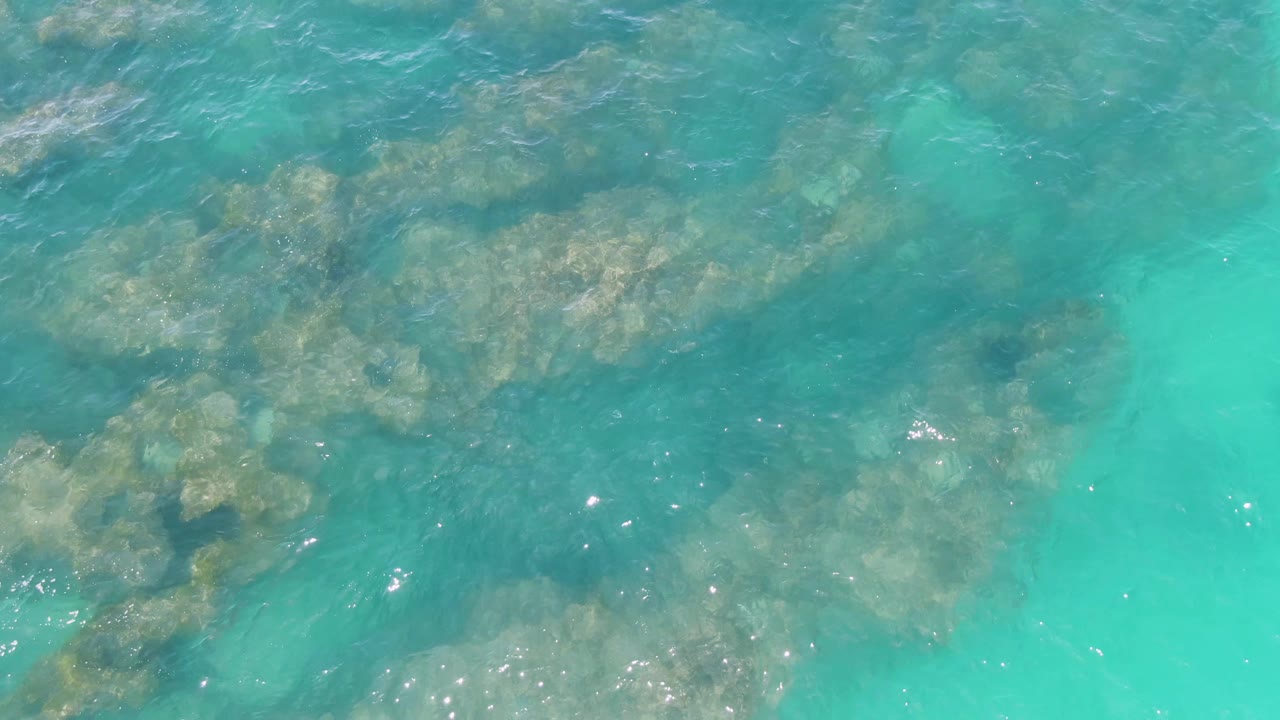 An overhead frame from Buzzards Beach shows submerged rock formations under turquoise water, with brighter aqua water along the right side and no visible people.