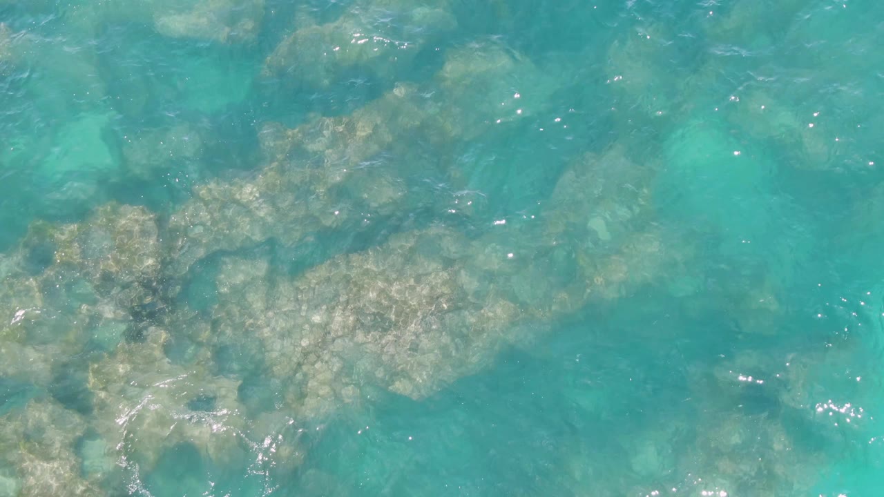 Clear blue-green seawater at Buzzards Beach, Baja California Sur, passes over elongated submerged rock formations, with bright glints near the right edge and no visible people.