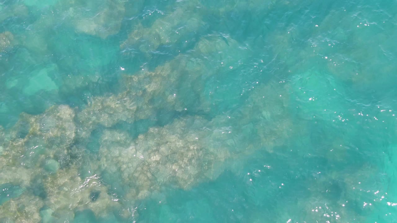 Shallow turquoise sea at Buzzards Beach, Baja California Sur, shows denser rock patches near the lower half of frame, faint sandy color through the water, and no visible people.