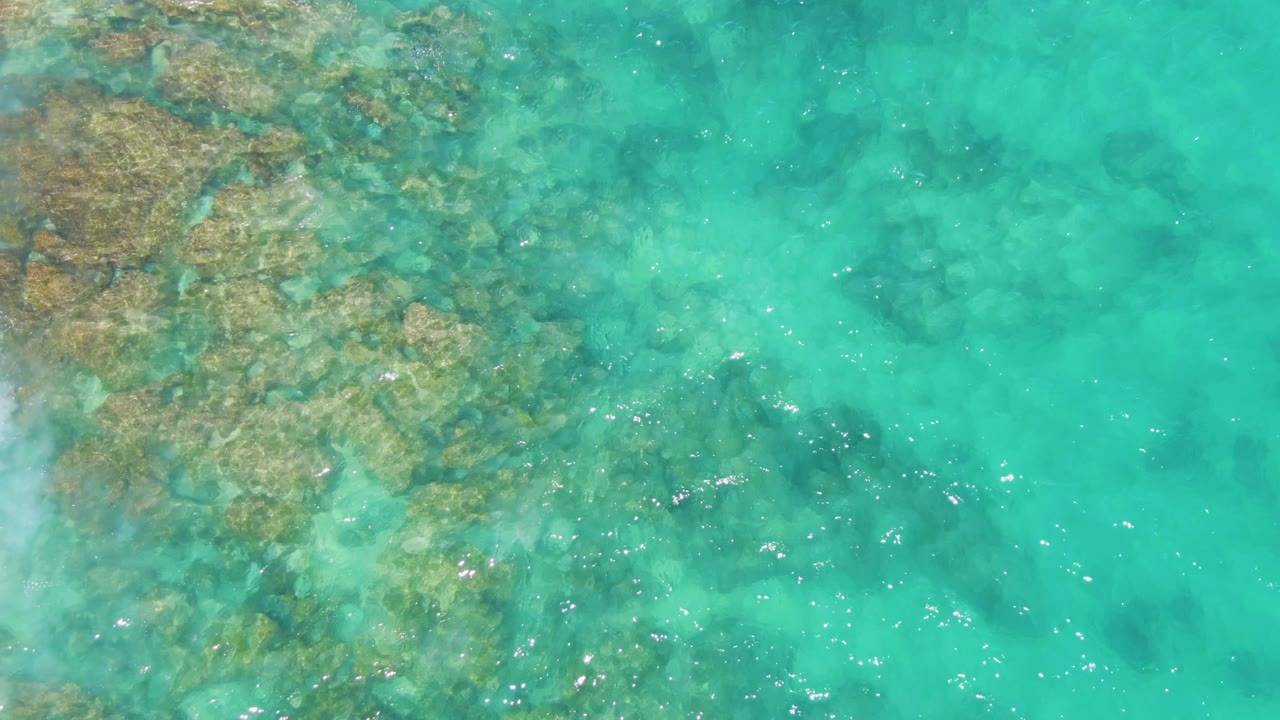 Downward aerial view of clear turquoise water over submerged rocks at Buzzards Beach, Baja California Sur, with small sun glints and no visible people.
