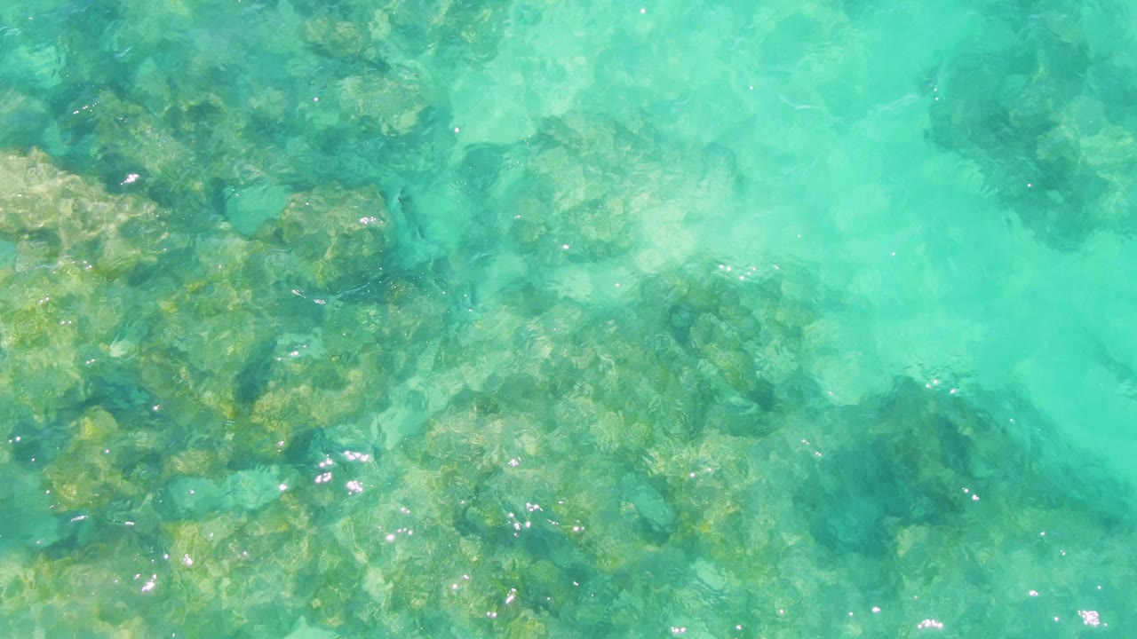 Top-down view of turquoise water over submerged rocks at Buzzards Beach in Baja California Sur, with soft ripples and no visible people.