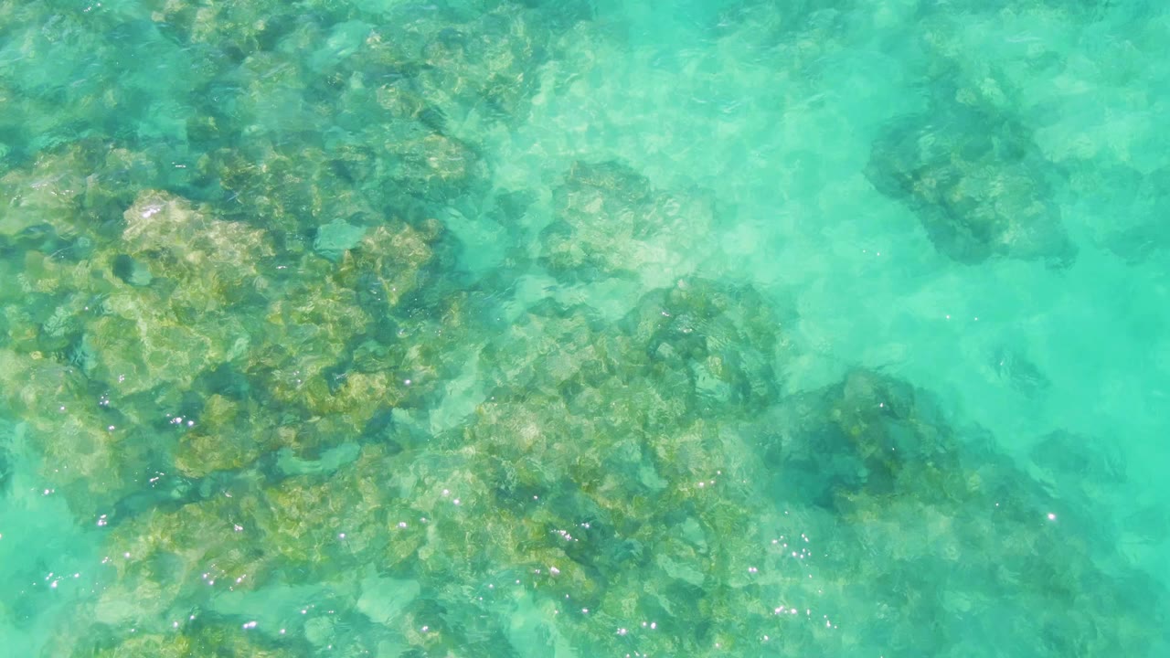 Turquoise water at Buzzards Beach covers clustered underwater rocks, with a lighter sandy-looking patch near the center and no visible people.
