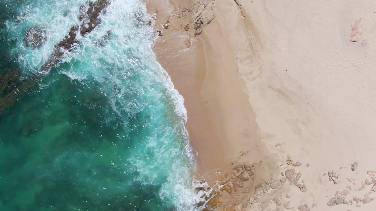 Top-down view of turquoise water and white foam reaching the sandy edge of Buzzards Beach, Baja California Sur, with dark rocks in the surf and no visible people.