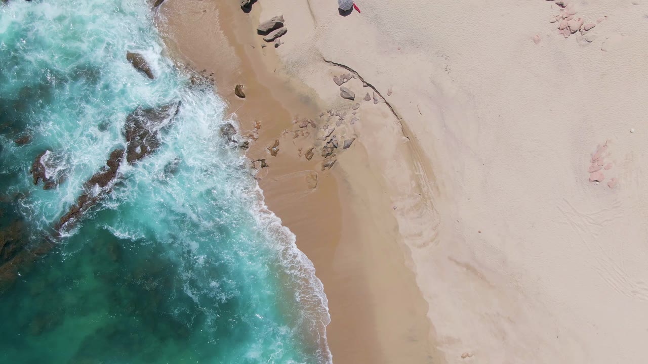 Overhead Buzzards Beach view with white surf curling around several dark rocks and smooth sand extending across the right side, with no visible people.