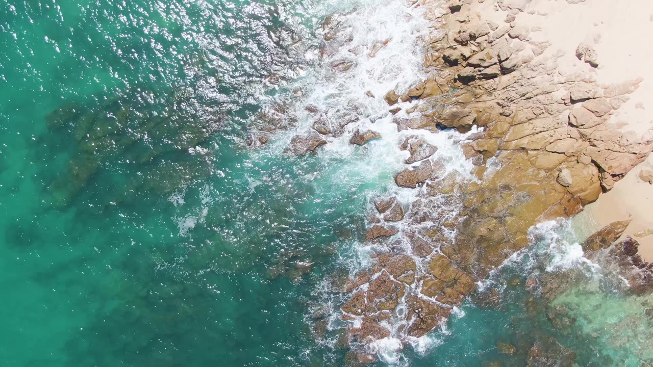 Aerial frame over Buzzards Beach with white surf spreading across tan rocks, darker underwater stones in turquoise water, and pale sand at the upper right, with no visible people.