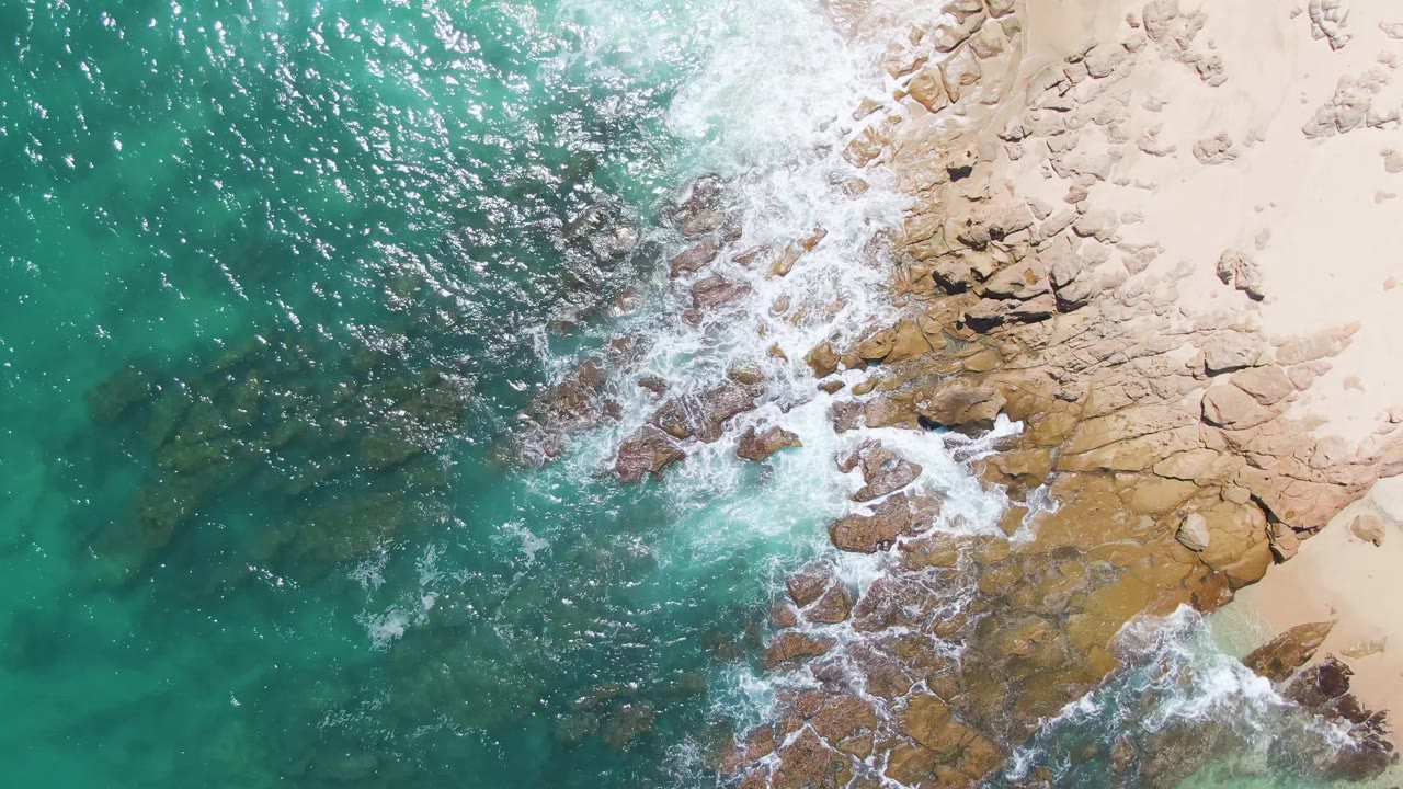 Top-down Buzzards Beach frame showing green-blue ocean on the left, white water around the rocks, and a larger area of pale sand along the upper right edge, with no visible people.