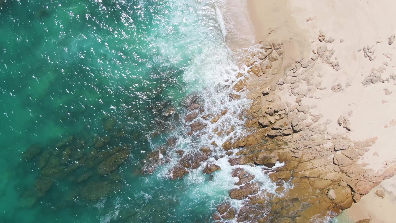 Aerial view at Buzzards Beach with clear turquoise water filling the left frame, white foam crossing a rocky point, and pale sand with scattered rocks on the right, with no visible people.