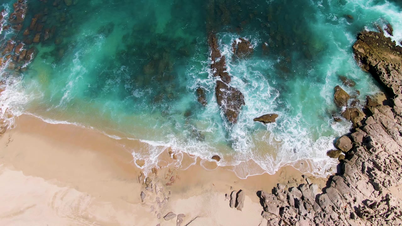 Overhead turquoise surf, tan sand, and dark rocks along a beach