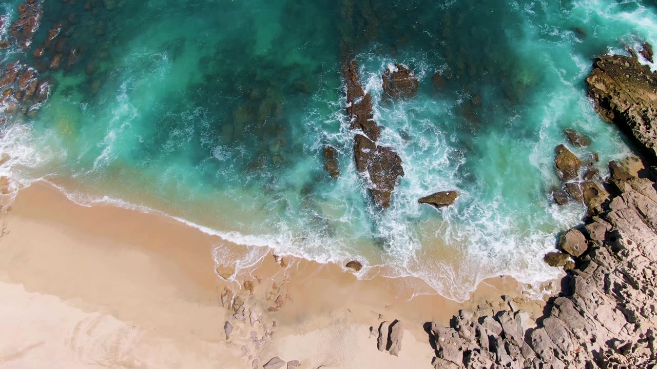Aerial frame of white foam spreading across turquoise water and pale sand at Buzzards Beach, with the main rock formation shifted toward the right and no visible people.