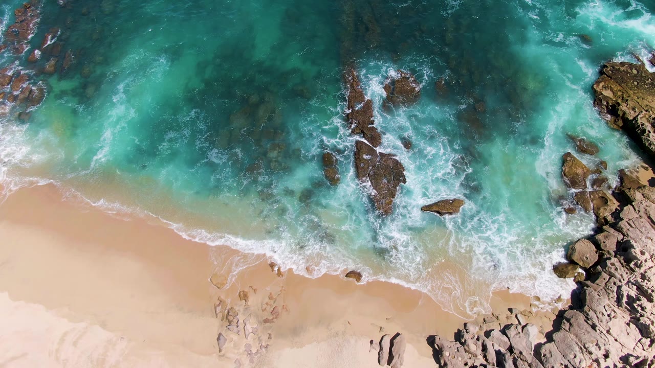 Top-down drone view of Buzzards Beach surf with darker submerged rock patches in the water, a rocky point at the right edge, tan sand below, and no visible people.