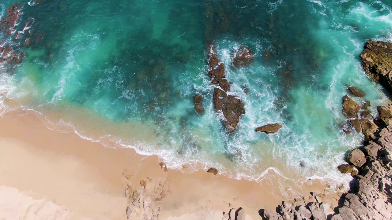 Overhead view at Buzzards Beach where white water breaks along the sand and around clustered brown rocks, with no visible people or structures.