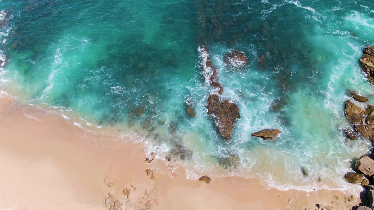 Overhead view at Buzzards Beach shows turquoise water breaking around dark rocks near pale sand, with no visible people.