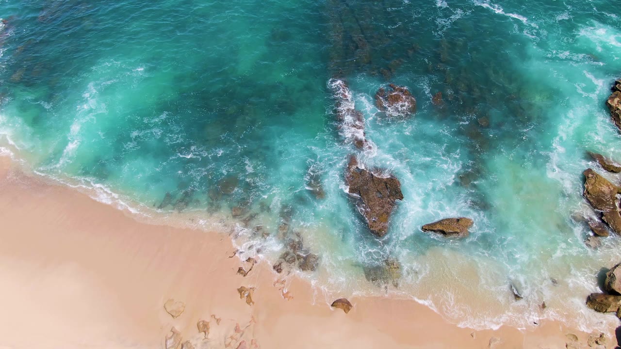 At 0:01, a white foam line curls across the sand at Buzzards Beach while several rocks sit in shallow turquoise water, with no visible people.