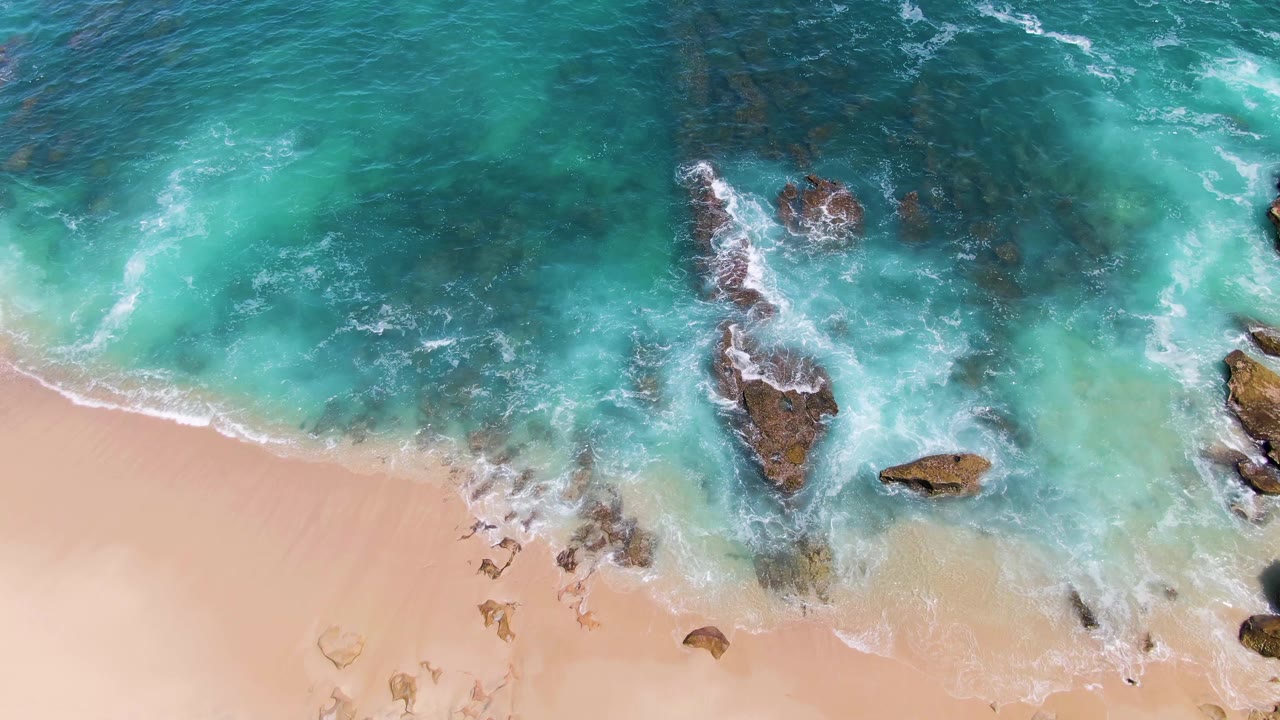 At 0:02, clear blue-green water covers submerged rocks as foam breaks along the Buzzards Beach shoreline, with no visible people.