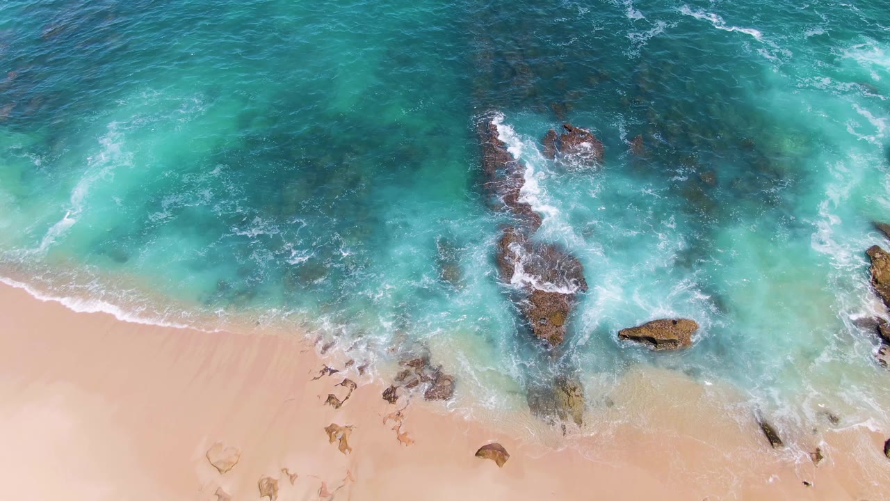 At 0:03, the overhead composition places pale beach along the lower frame and foamy turquoise surf around a vertical rock formation at Buzzards Beach, with no visible people.