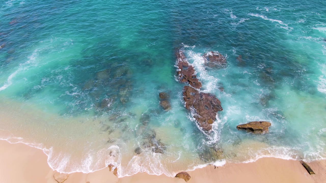 Aerial frame from Buzzards Beach showing white foam curling across the lower sandy edge while clear blue-green water surrounds exposed rocks, no visible people.