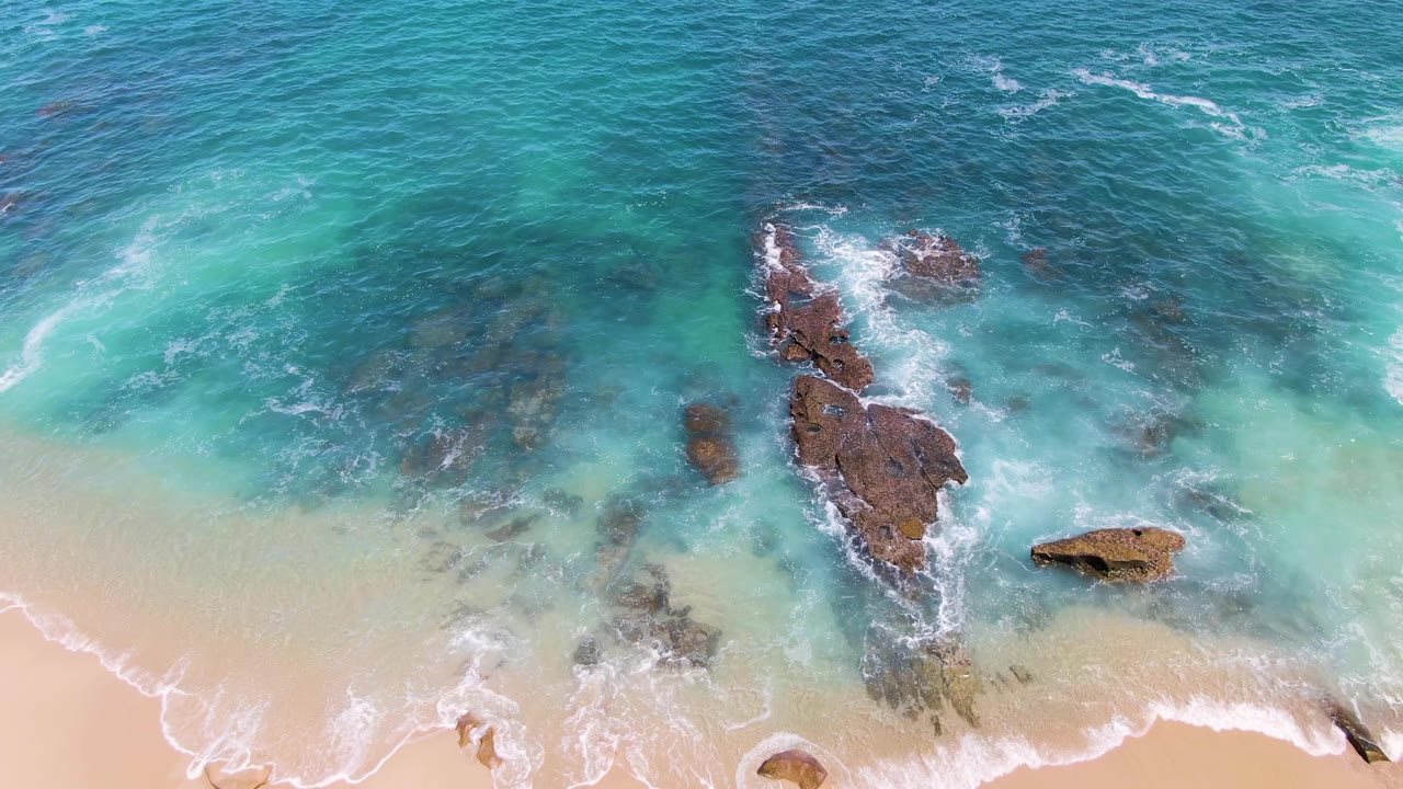 Top-down Buzzards Beach view with a larger dark rock cluster near the right center, pale sand below, and foam streaks moving through turquoise water, no visible people.
